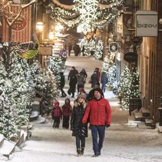 two people walking down a snowy street in front of christmas trees and lights on the buildings