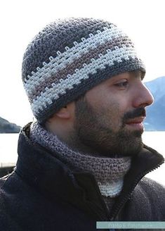 a man with a beard wearing a knitted hat next to the water and mountains