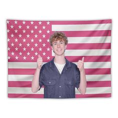 a young man giving the thumbs up sign in front of an american flag wall tapestry