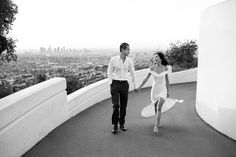 a man and woman holding hands walking down a hill with the city in the background