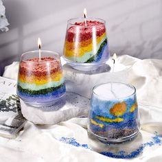 three candles are sitting on a table with some sand in them and one has a rainbow painted candle