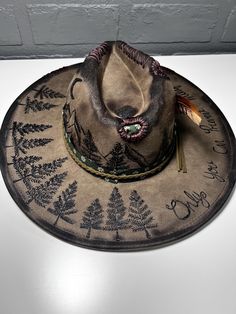 A hand-burned wide-brim hat with tree designs, mountain details, a feather accent, and "Only You can prevent forrest fires" written on the brim. Burned Designs On Hats, Custom Burned Hat Ideas, Rustic Curved Brim Hunting Hat, Custom Brimmed Fedora For Outdoor, Custom Handmade Hats For Outdoors, Rustic Flat Brim Felt Hat For Outdoor, Rustic Felt Hat With Flat Brim For Outdoor, Rustic Brimmed Felt Hat For Outdoor, Bohemian Short Brim Felt Hat For Outdoor