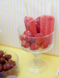 some strawberries and grapes in a glass bowl on a yellow table with other fruit