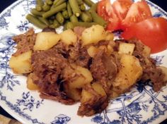 a blue and white plate topped with meat, potatoes and green beans next to tomatoes