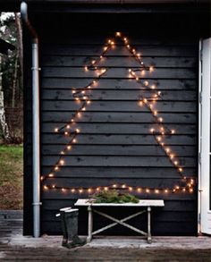 a christmas tree made out of lights on the side of a black building with a bench in front of it