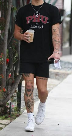 a man walking down the street with tattoos on his legs and leg, holding a drink