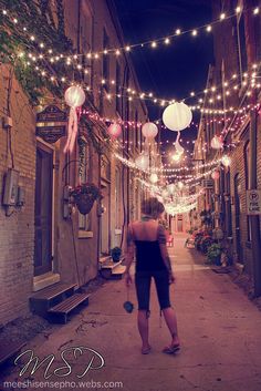 a woman is walking down an alley way with lights strung all over the street and buildings