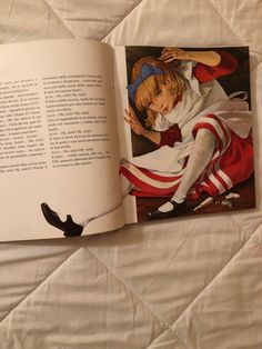 an open children's book on a bed with white sheets and red striped dress