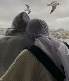 two people are looking at the seagulls flying in the sky above them and one person is wearing a white hoodie