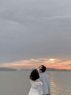 a man and woman standing next to each other in front of the ocean at sunset