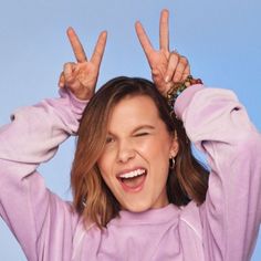 a woman making the peace sign with her hands while wearing a purple shirt and smiling