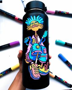 a person holding up a black water bottle with colorful drawings on it and crayons in the background