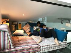 two people are sitting on a couch with pillows and books in their hands while reading