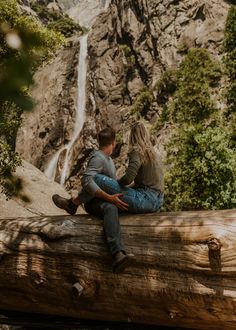 a man and woman sitting on a log in front of a waterfall while looking at each other