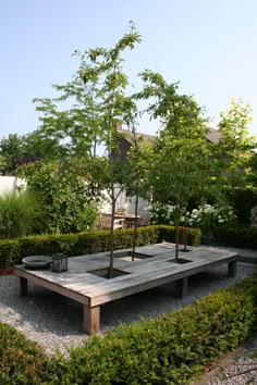 a wooden table surrounded by bushes and trees in a garden area with graveled ground