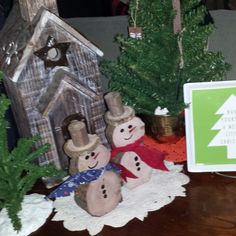 there are snowmen standing in front of a small house and christmas trees on the table