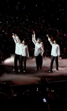 three men in white shirts and black pants standing on stage with their hands up to the sky