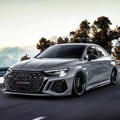 the front end of a silver car driving on a road with trees in the background