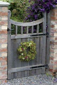 a wooden gate with a wreath hanging on it's side and purple flowers in the background