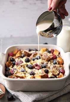 someone pouring milk into a casserole dish with berries and bread on the side