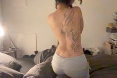 a woman with a tattoo on her back standing in front of a bed and looking at the camera