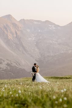 a bride and groom standing on top of a grass covered hillside with mountains in the background