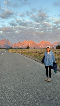 a woman is standing on the side of an empty road with mountains in the background