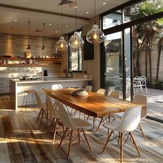 a dining room table with chairs and lights hanging from it's ceiling, in front of an open kitchen