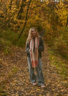 a woman with long hair standing on a path in the woods wearing jeans and a scarf