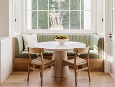 a white table sitting under a window next to a green bench with two wooden chairs