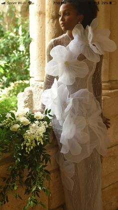 a woman standing next to a wall with flowers in her hair and wearing a white dress