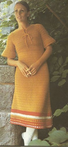 a woman standing next to a stone wall wearing an orange crocheted dress and white socks