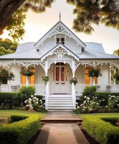 a white house that is surrounded by bushes and trees with flowers on the front door