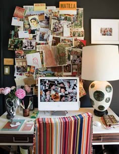 a desk topped with a computer next to a lamp and pictures on the wall behind it