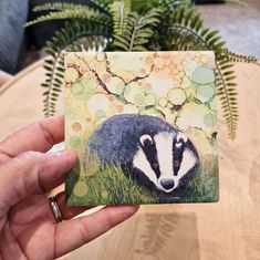 a hand holding up a small coaster with a badger painting on it's side