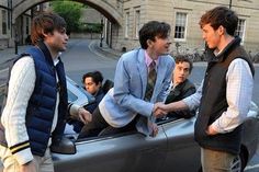 three men in suits are shaking hands over the top of a car while another man looks on