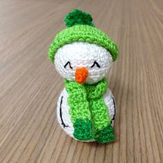 a small crocheted snowman sitting on top of a wooden table wearing a green hat and scarf