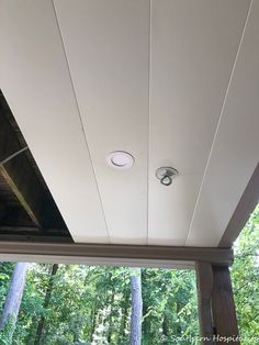 an outdoor covered porch with two lights on the ceiling and trees in the back ground