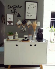 a white cabinet with some coffee cups and pictures on the wall next to it in front of a window