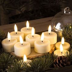 many white candles are lit on a tray with pine cones and greenery around it