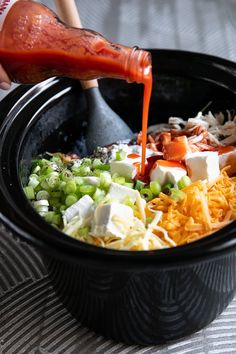 someone pouring dressing into a black bowl filled with different types of vegetables and cheeses