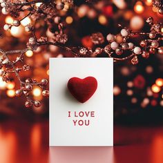 a card with a red heart on it sitting in front of a christmas tree and lights