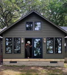 a woman standing in the open door of a small gray house with trees behind her