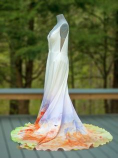 a white dress with orange and blue paint on it is hanging from a string in front of some trees