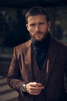 a man wearing a brown suit and black turtle neck sweater is looking at the camera