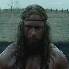 a man with long hair and beard standing in front of a body of water wearing a chain around his neck