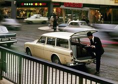 a man standing next to an old car on the side of a road with traffic in the background