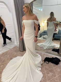 a woman standing in front of a mirror wearing a white wedding dress with off the shoulder sleeves