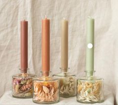 four candles are lined up in glass containers with dried flowers and leaves inside them on a white cloth