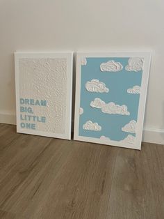 two framed art pieces on the floor in front of a white wall with blue clouds